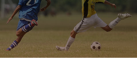 Two soccer players competing on a grass field, with one player kicking a soccer ball while another runs alongside.